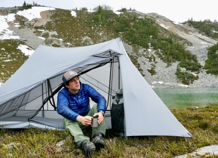 一名男子坐在帐篷里喝着咖啡，背景是山脉。