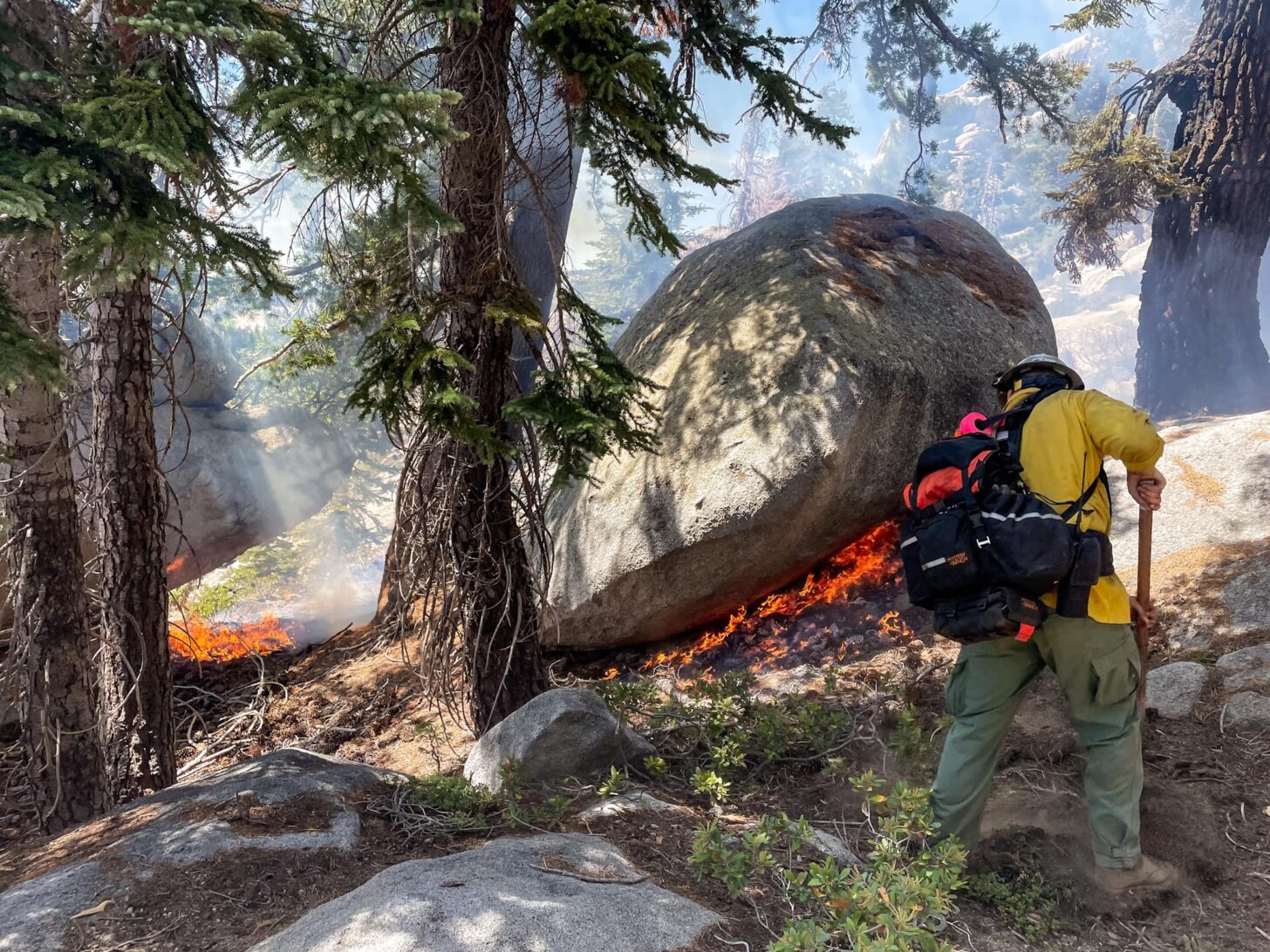 一名背着背包、戴着头盔的消防员正在挖掘地面。照片中烟雾模糊，中间的一块巨石下可以看到火焰。