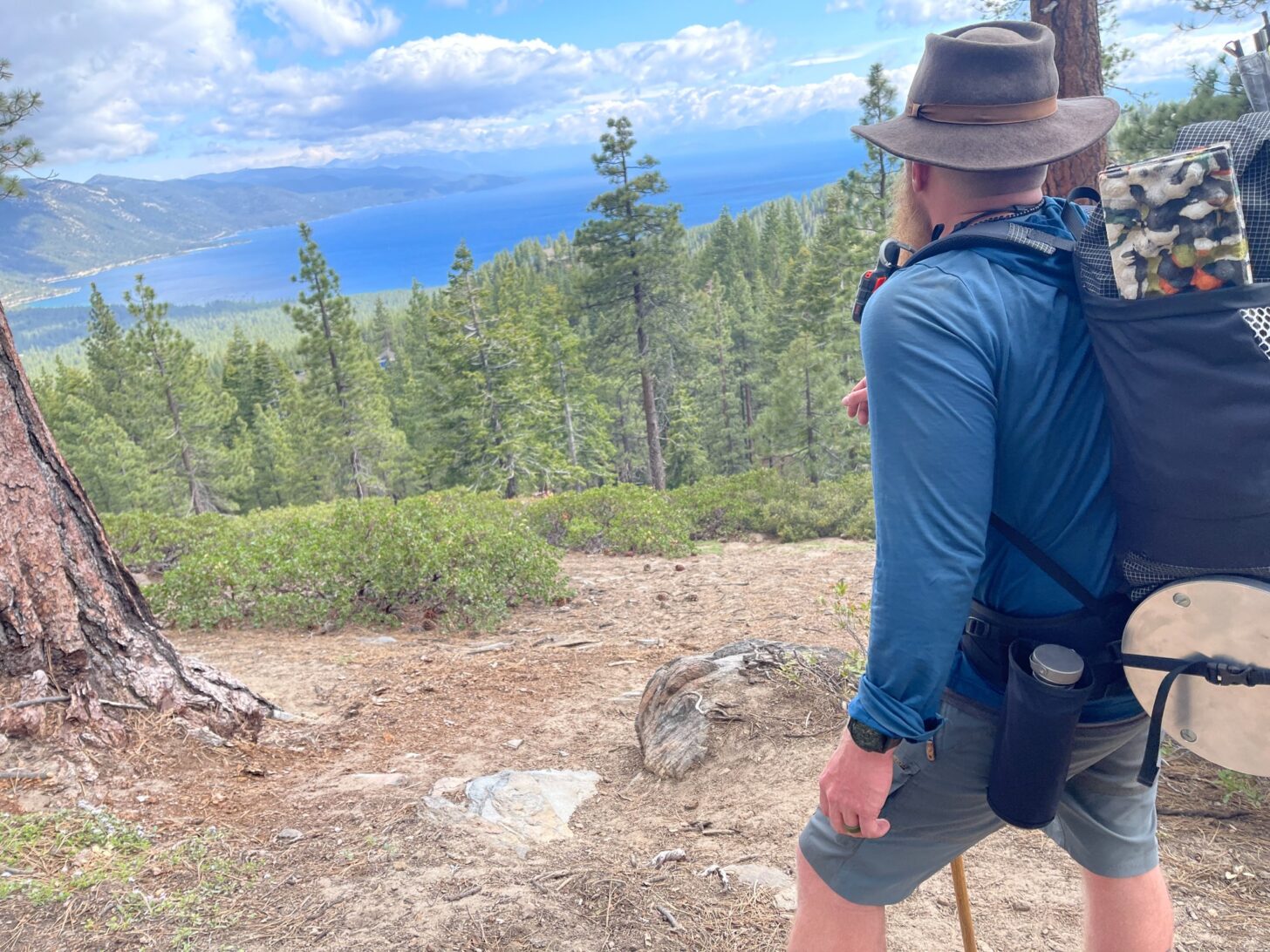 一个背着背包的男人站在那里，眺望着蓝色的湖泊和树木。在他腰部的一个小袋子里可以看到瓦戈瓶。
