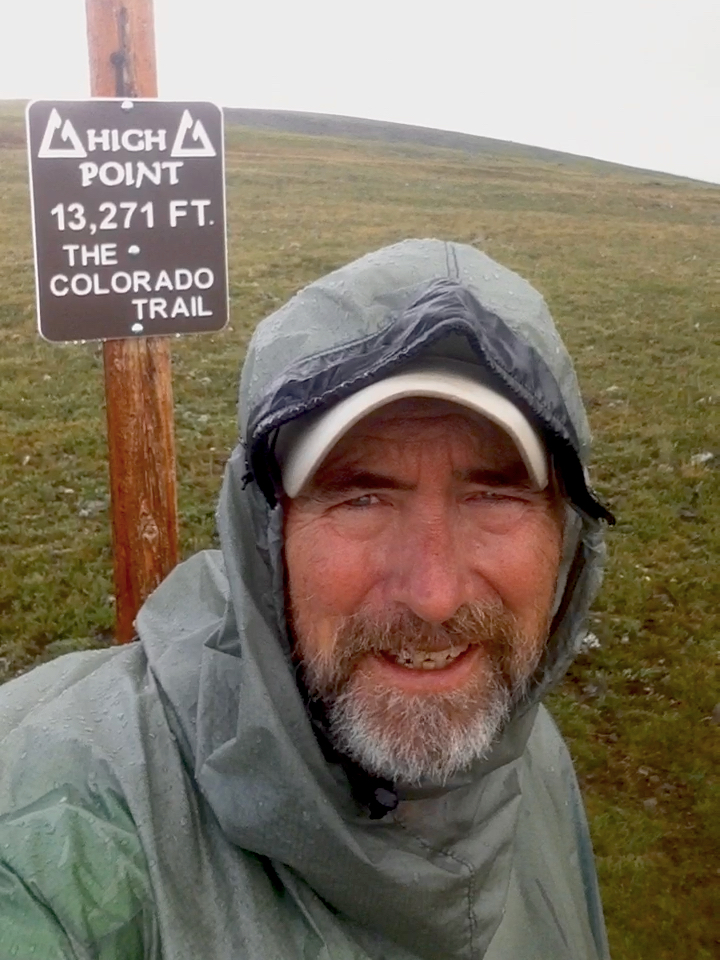 一名穿着雨披的男子在指示牌旁自拍。标志牌上写着“科罗拉多小径海拔13271英尺的高点”。