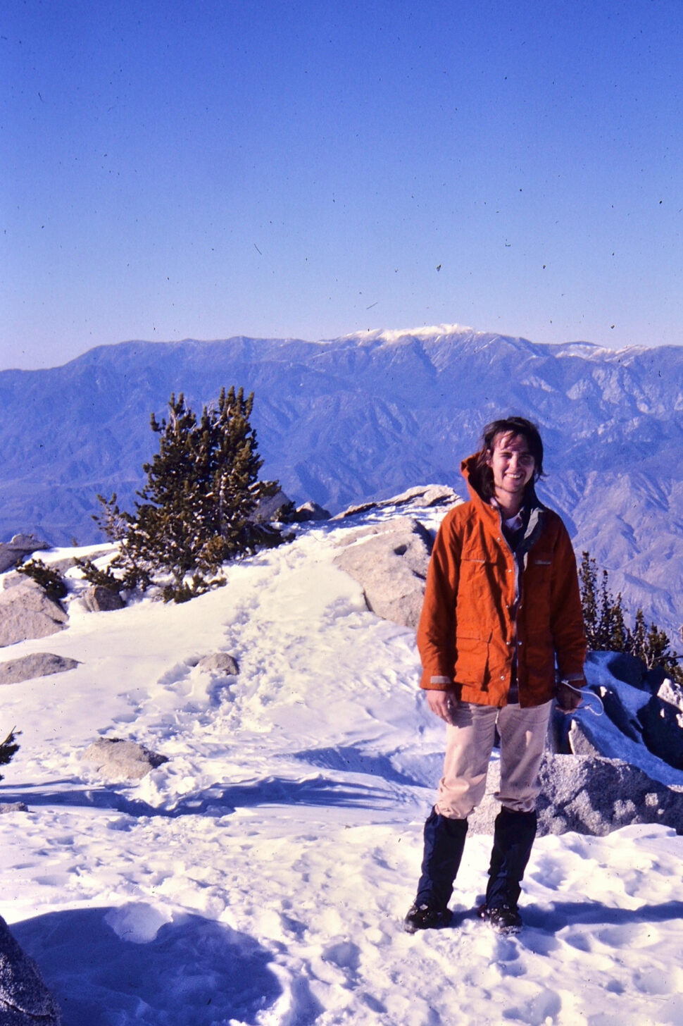 一名身穿橙色夹克的年轻人站在以群山为背景的雪峰上。