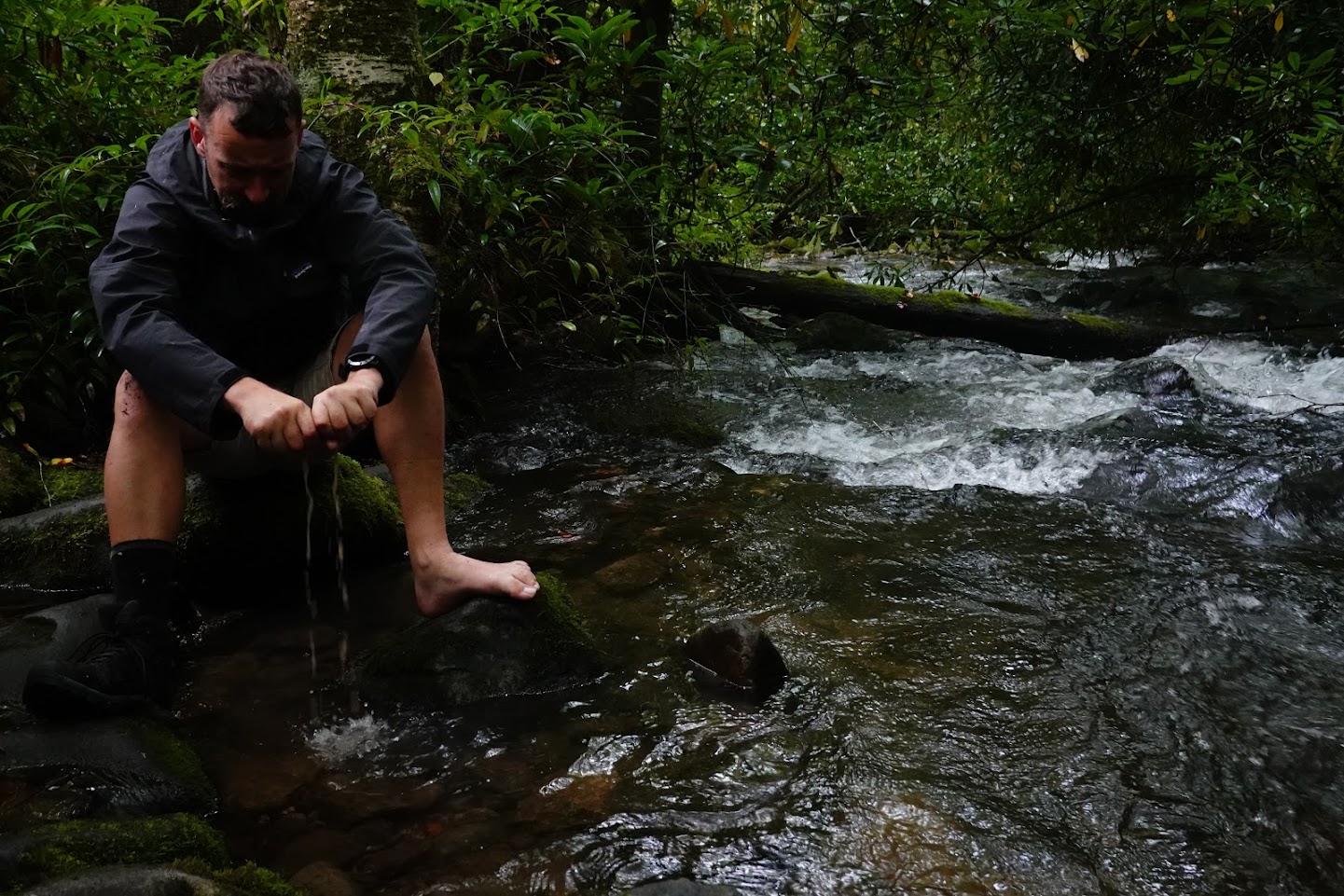 一名男子坐在小溪边的岩石上，挤出一只湿袜子。