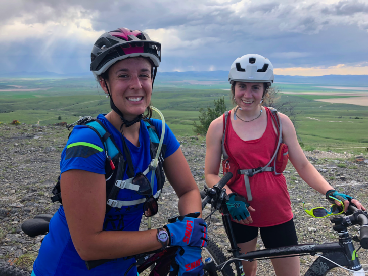 两名戴着自行车头盔站在自行车旁的女子对着镜头微笑。